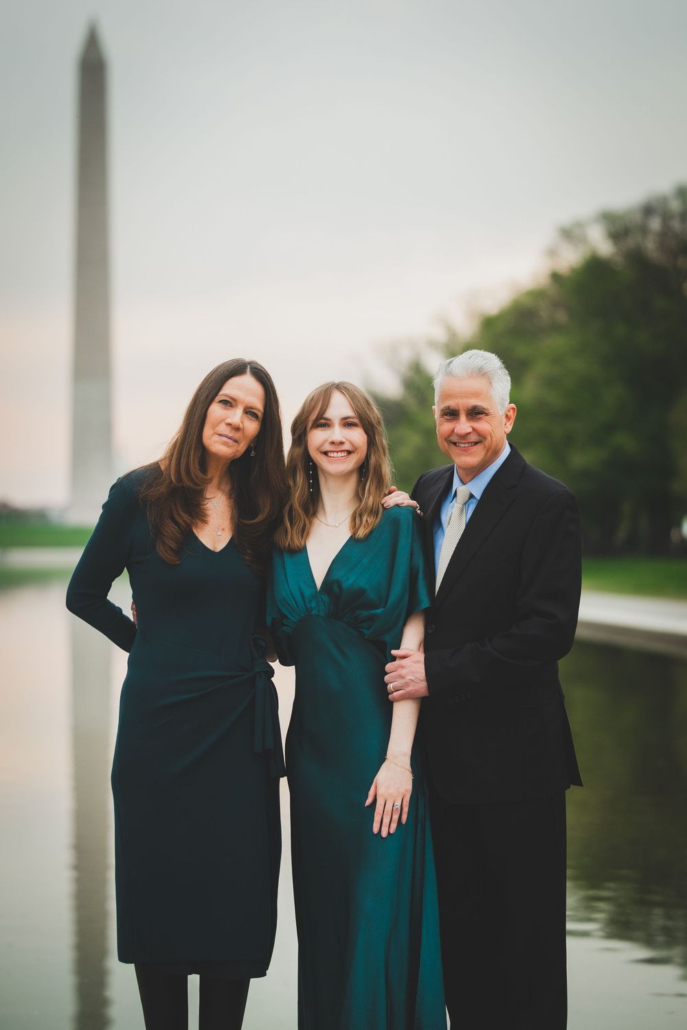 A family of three poses together outdoors near a reflective pool wearing coordinated dark formal attire.