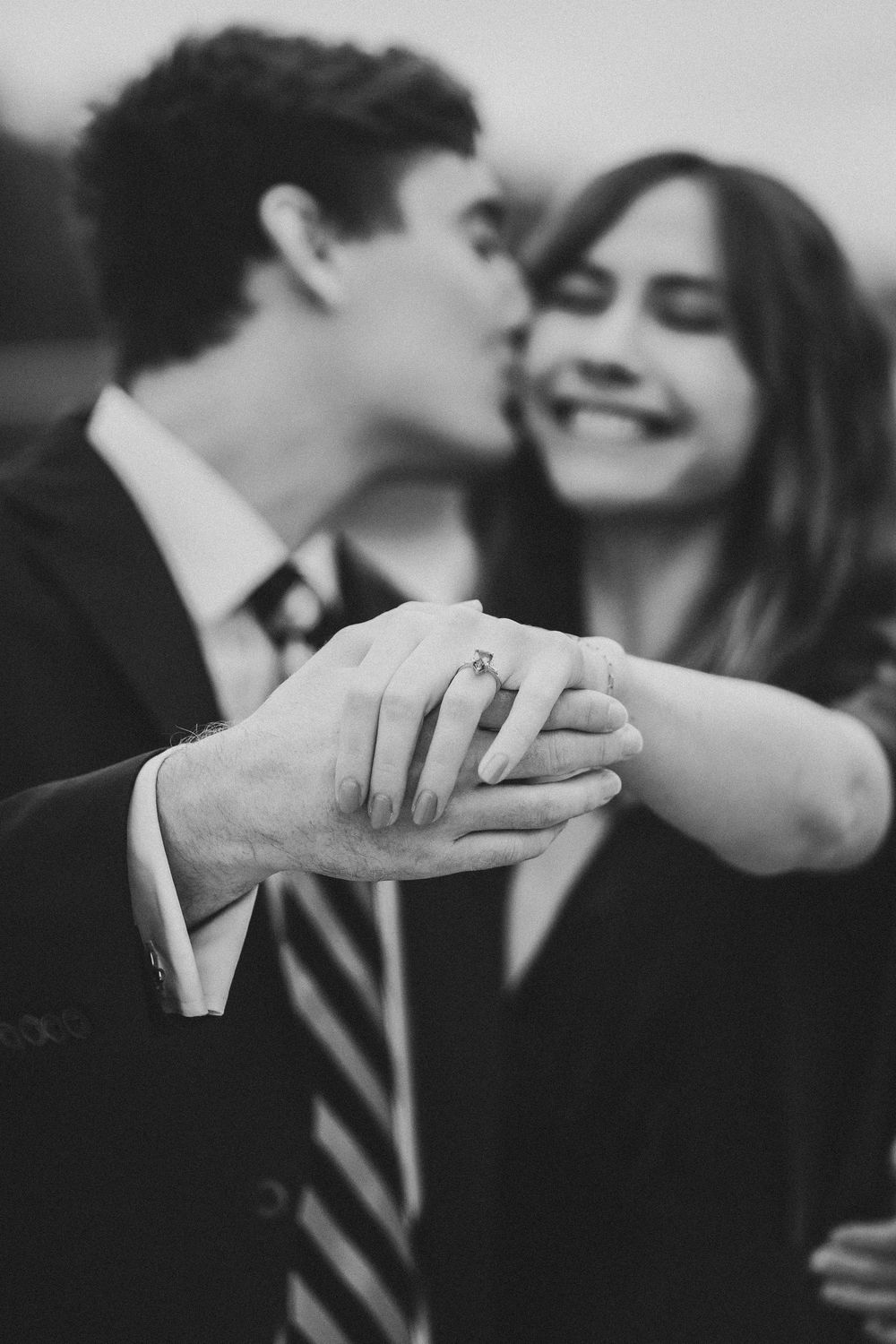 Black and white romantic engagement photo showing close up of hands with ring while couple shares tender moment.