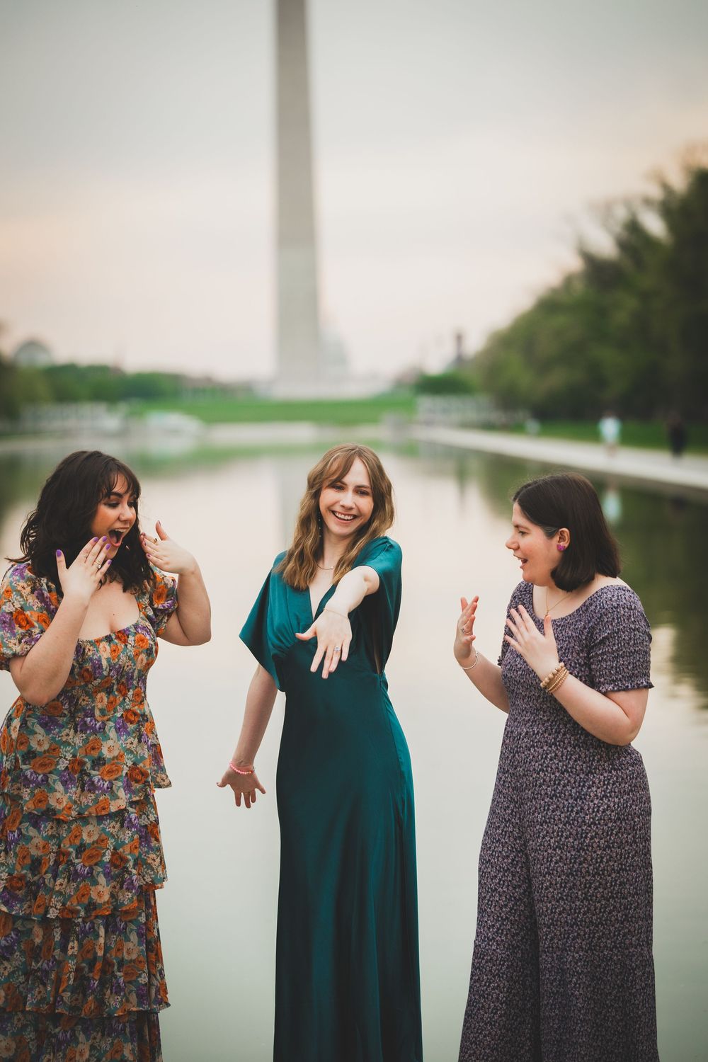 Three women in casual dresses share candid laughing moments by a reflecting pool in an outdoor photo series.
