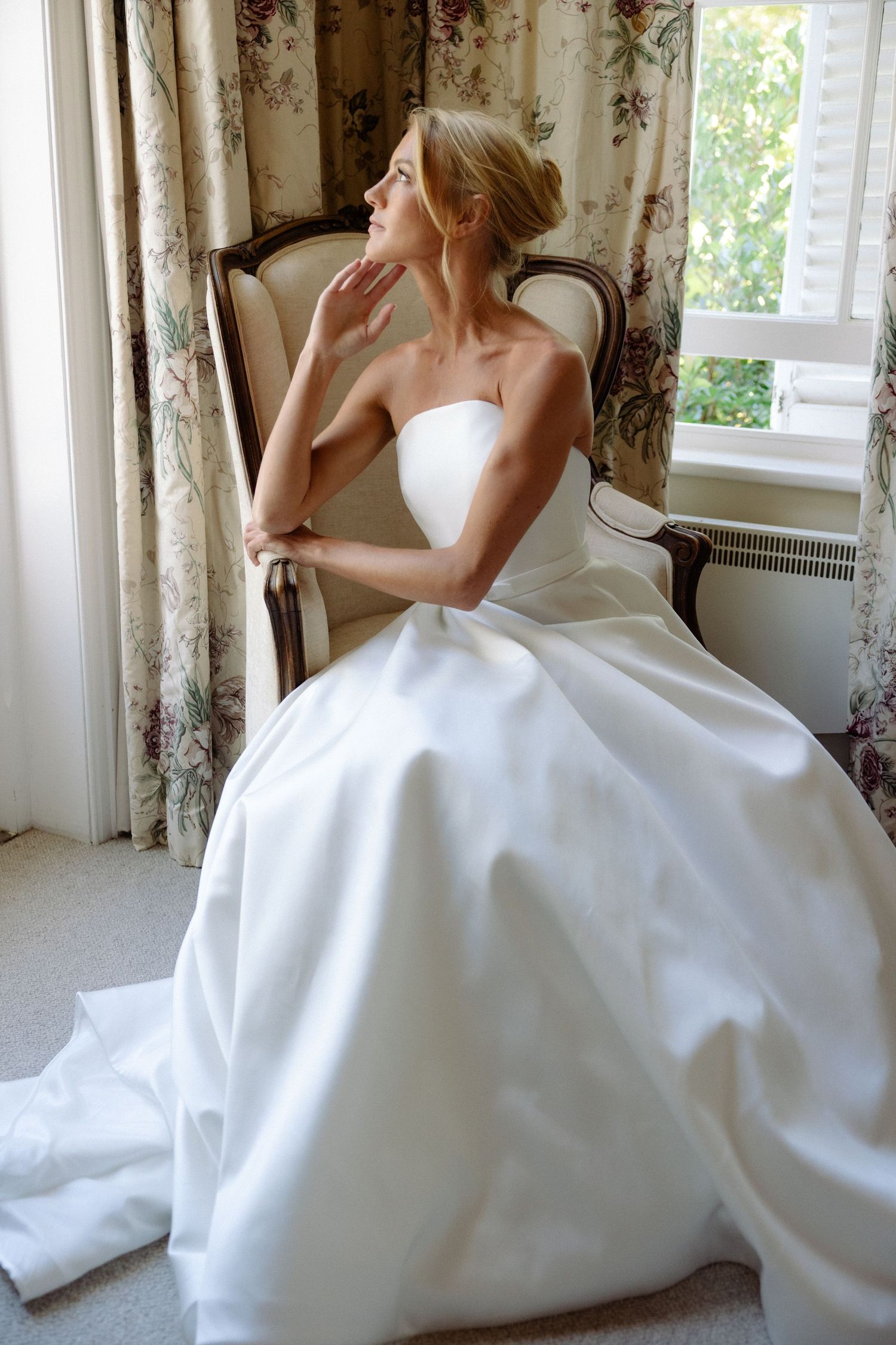 A person in a flowing white wedding gown sits gracefully by a window with floral curtains in a bright room.