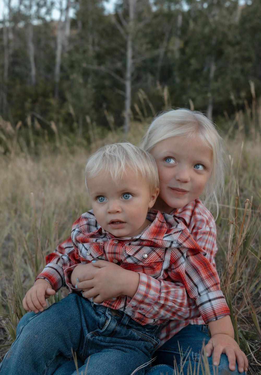Two young children in plaid shirts sitting together in tall grass with playful expressions.