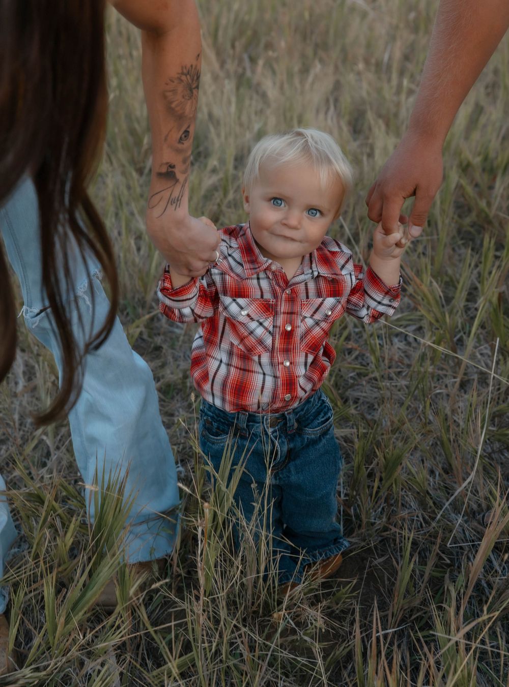 Young child in plaid shirt and jeans playing in a field of tall grass at sunset.