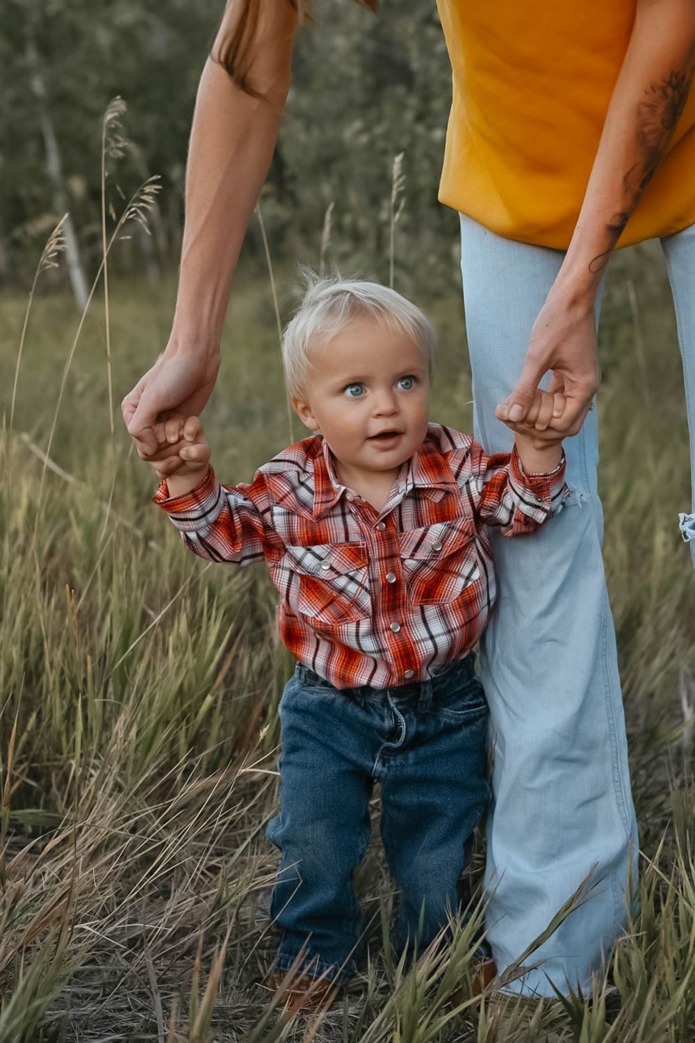 Small child in plaid shirt holds hands with adults while walking through tall grass.