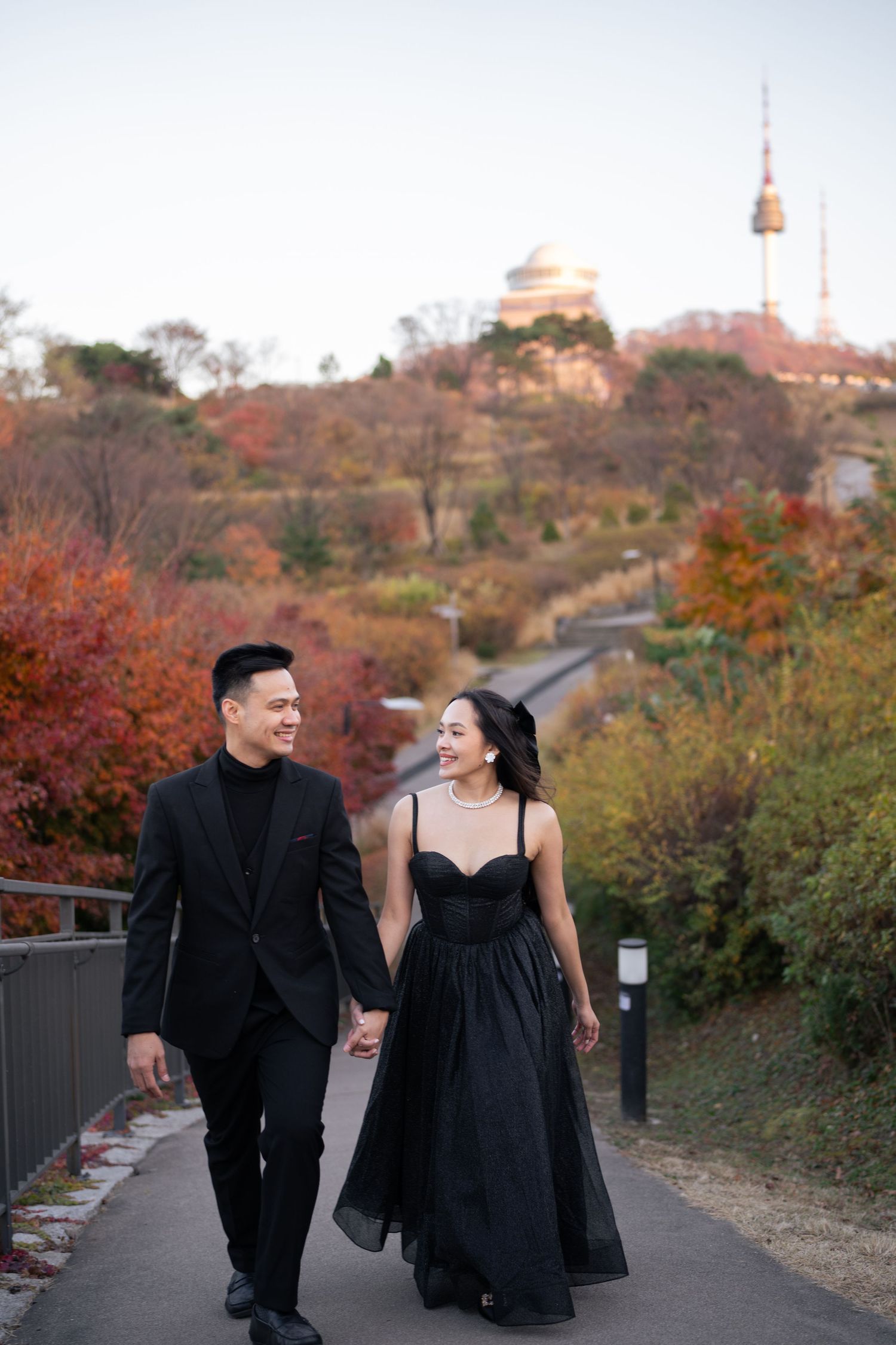 A couple in black formal attire walks and laughs together on a path during autumn with fall foliage in the background.