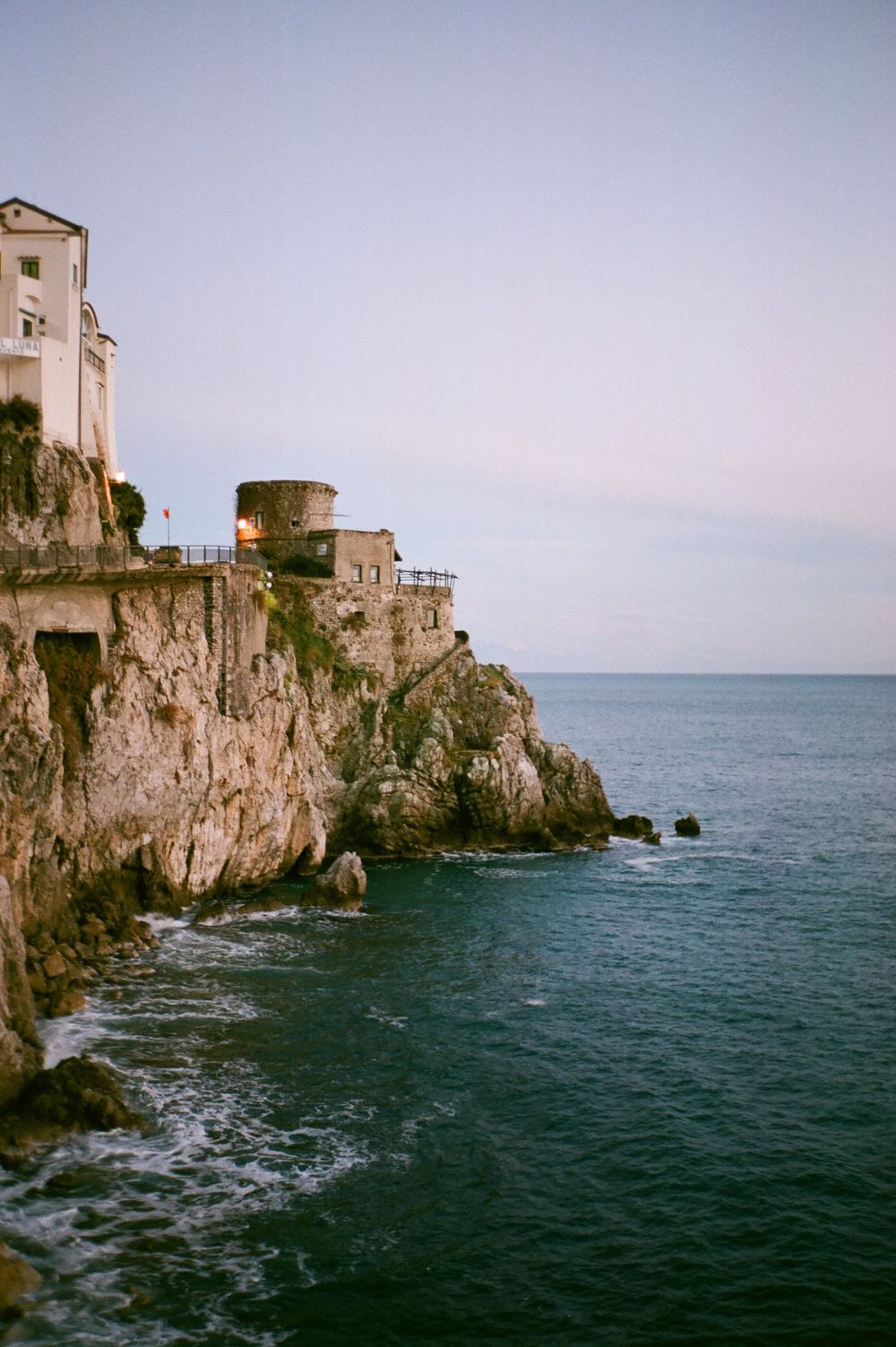Historic stone castle perched dramatically on coastal cliffs overlooking the deep blue Mediterranean Sea at sunset.