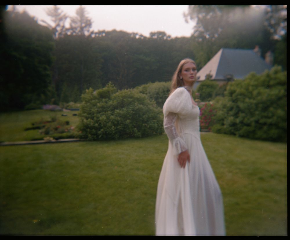A dreamy vintage-style photograph shows someone in a flowing white dress standing in a lush garden at dusk.