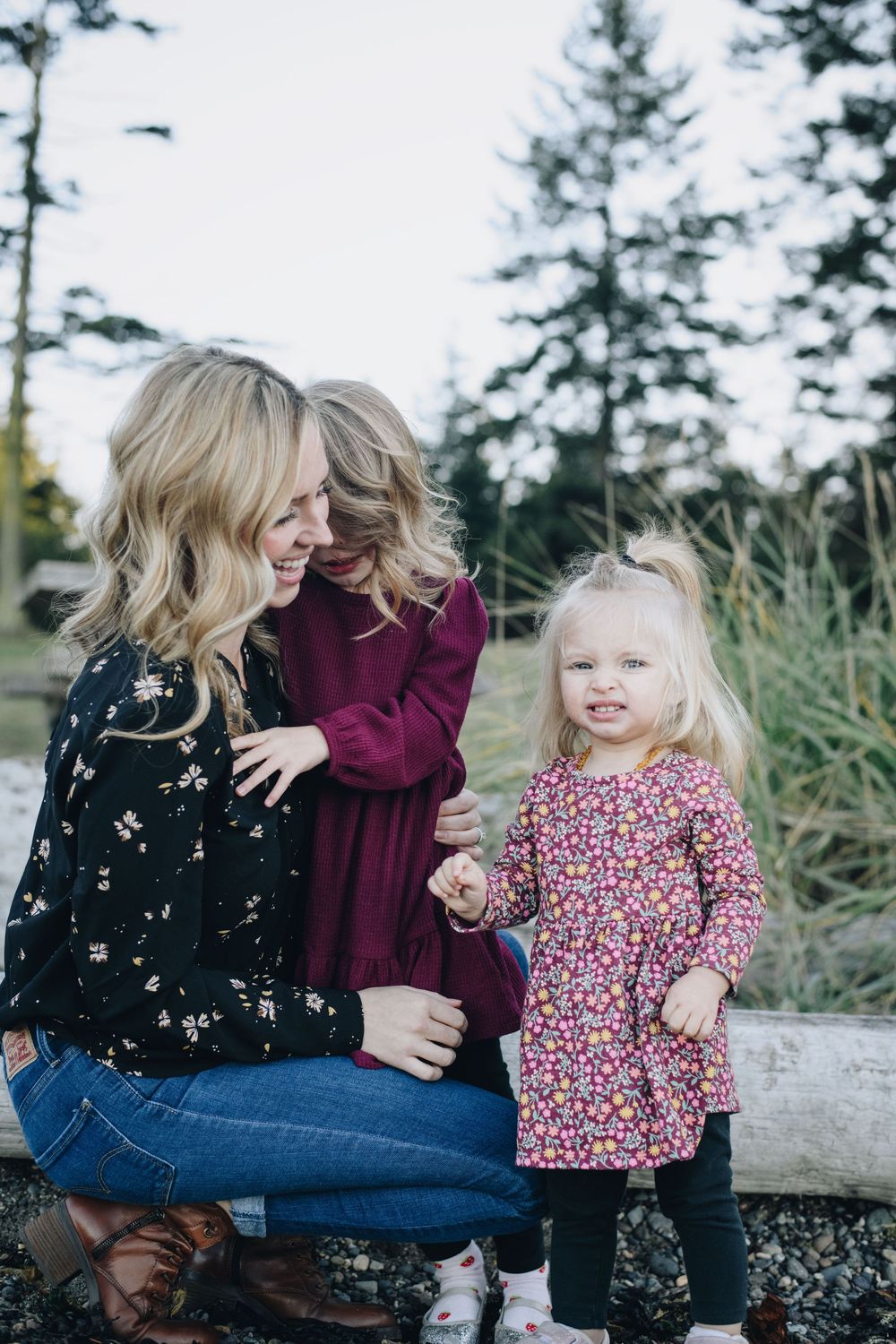 A casual outdoor family moment with three people laughing together while sitting on a log among evergreen trees.