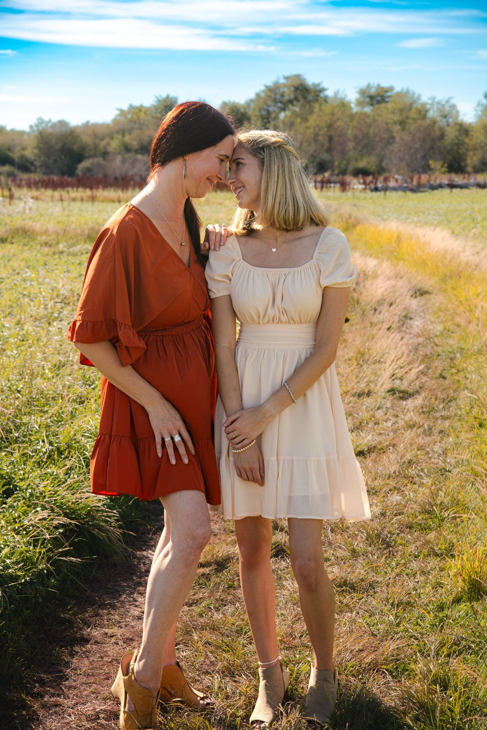 Two people in coordinating rust and white dresses stand together in a golden field at sunset.