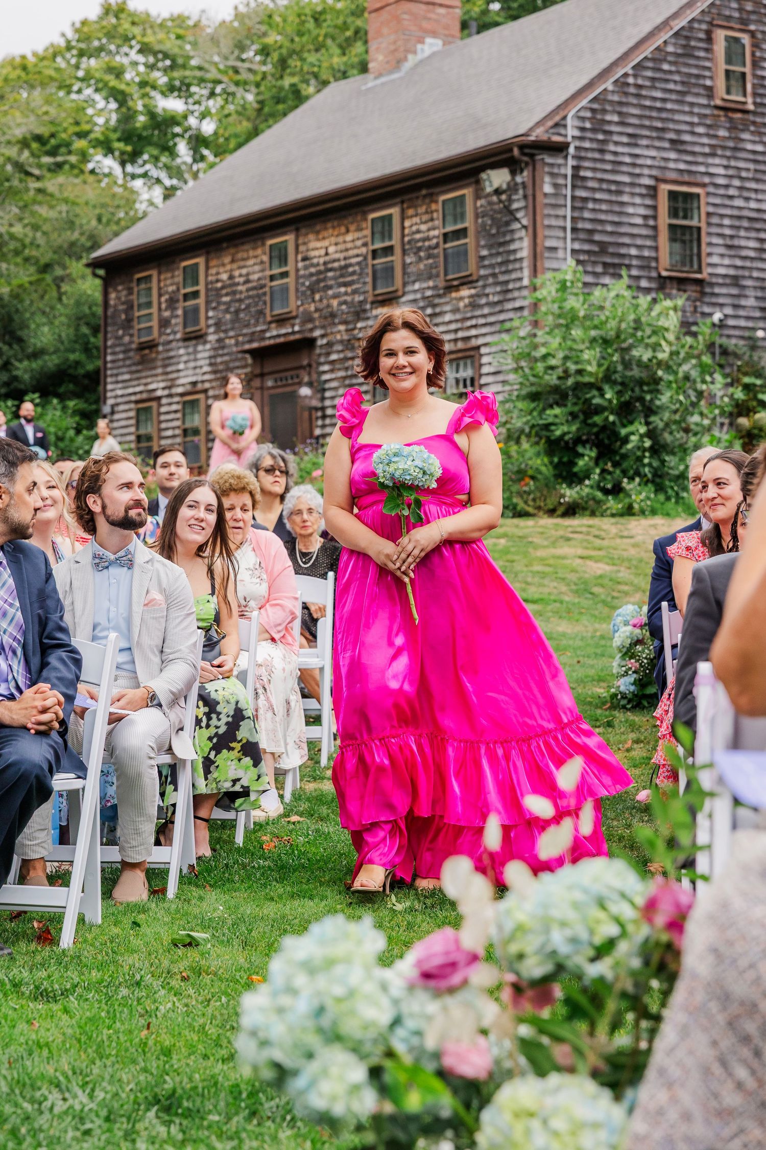 A wedding ceremony scene at a rustic barn venue with pink floral decorations and white chairs set up on a grassy lawn.