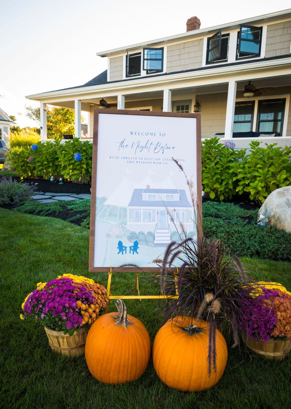 Decorative fall display with orange pumpkins, purple mums, and rustic signage arranged on a grassy lawn at dusk.