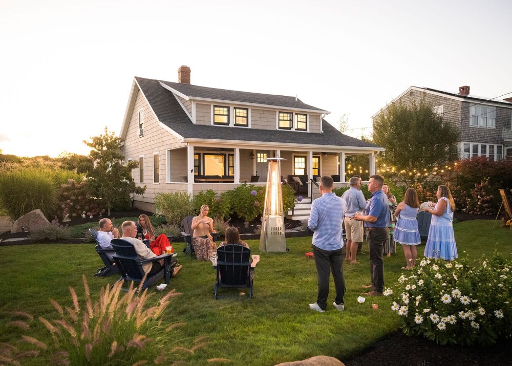 People gather on a grassy lawn at sunset with houses visible in the background during a social gathering.
