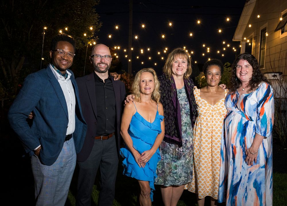 A group of people laughing and socializing at an evening event with warm lighting in the background.