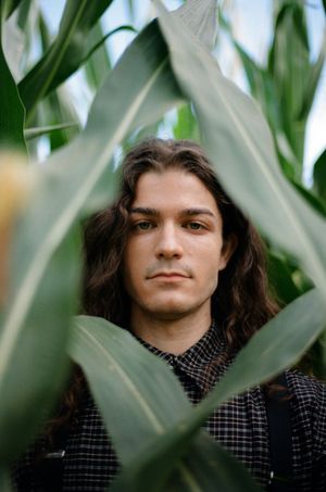 Portrait through green leaves showing someone wearing a checkered shirt with long dark hair.