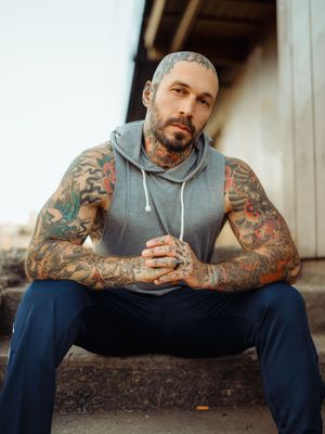 A person with colorful sleeve tattoos sits outdoors wearing a gray hoodie and navy pants.