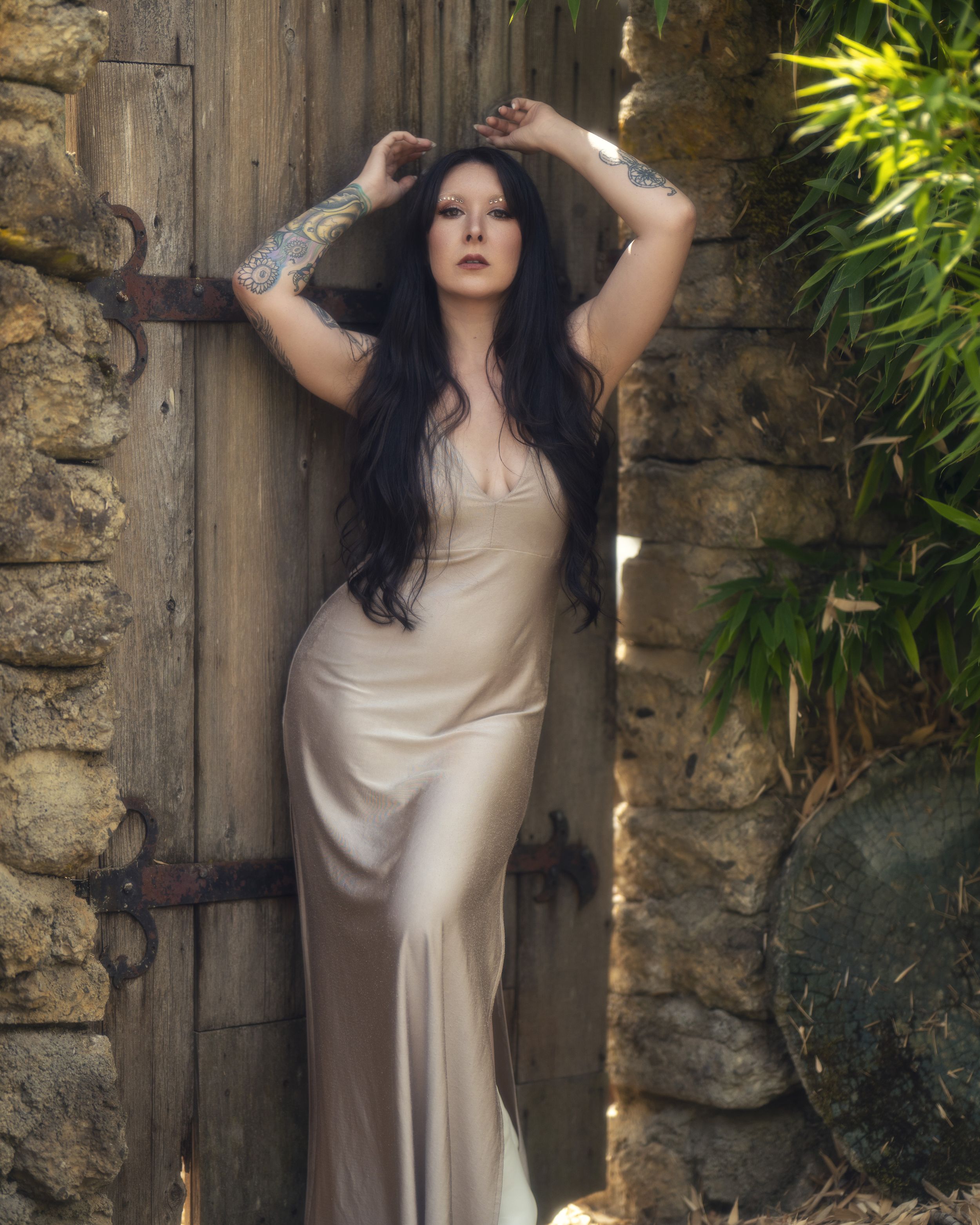 A person in a flowing cream-colored dress poses elegantly against a rustic stone wall and wooden door in natural light.
