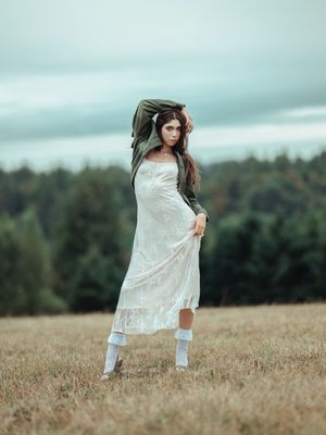 A figure in a flowing white dress stands gracefully in a golden field with trees in the distance on a misty day.