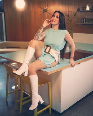 A person in a mint green top and white boots poses on a white bench in a warmly lit room.