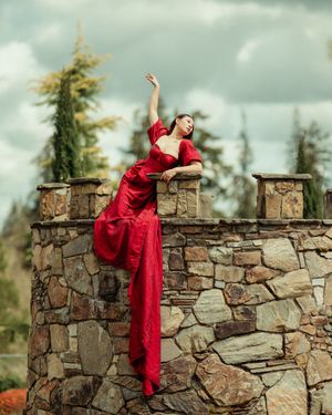 A person in a flowing red dress performs a graceful dance sequence on a rustic stone wall against a cloudy sky.