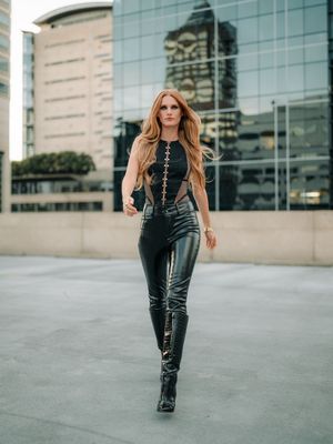 A person in black leather pants and boots poses confidently on an urban plaza with modern office buildings in the background.