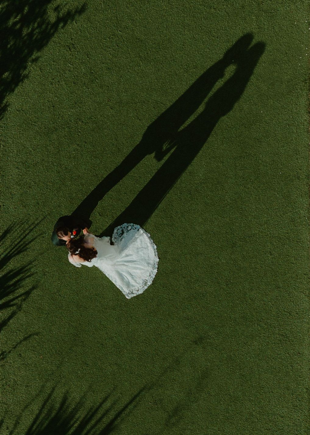 Aerial view of a couple in wedding attire lying on lush green grass creating long dramatic shadows.