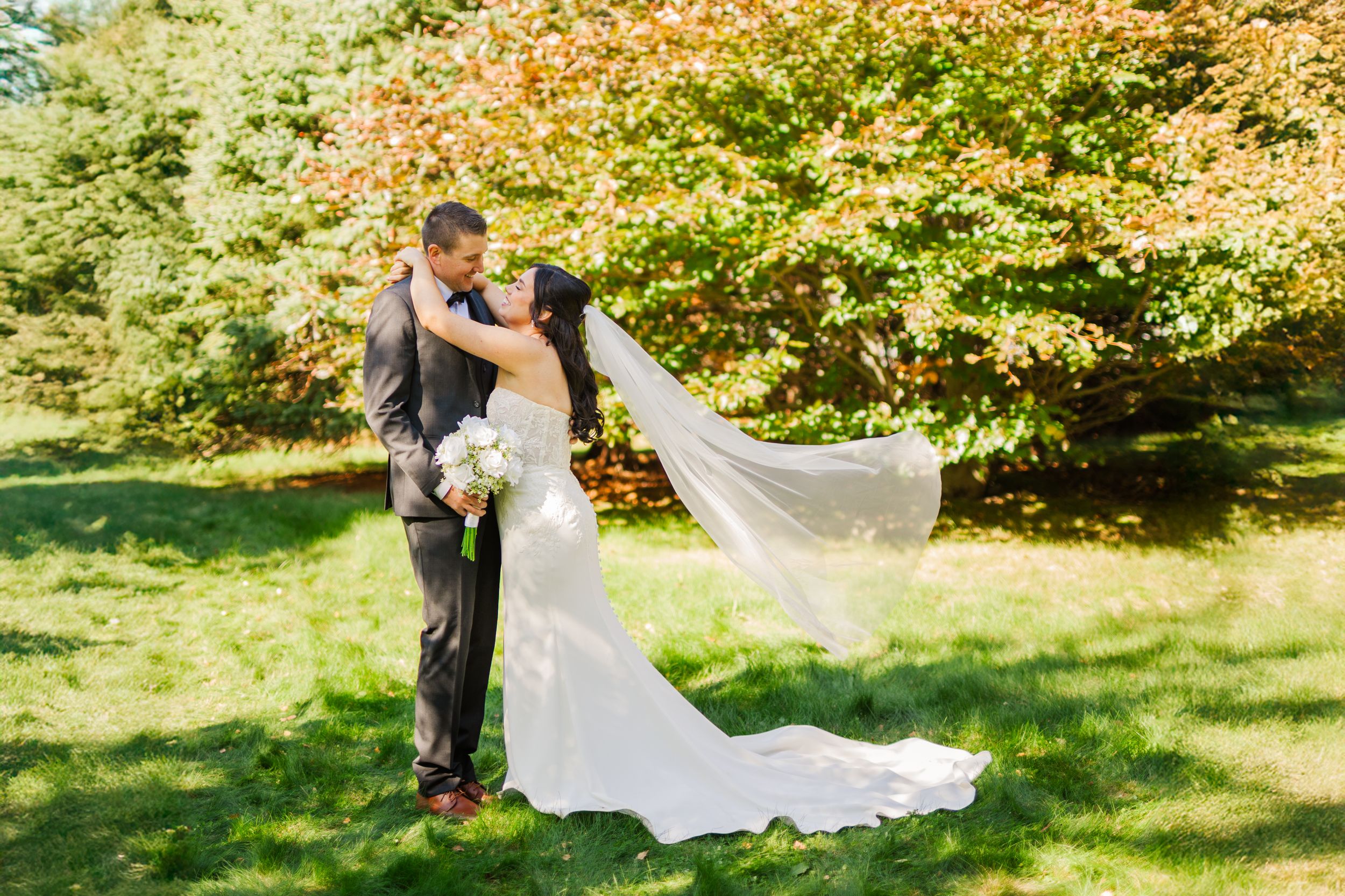 A couple in wedding attire embraces under sunlit autumn trees in a romantic outdoor setting.