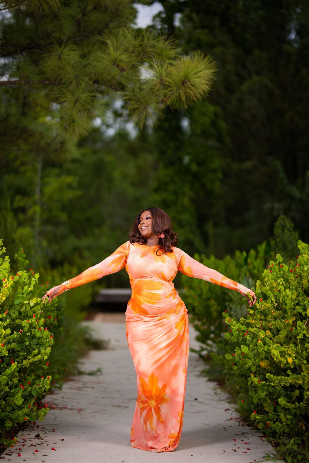 Figure in flowing orange dress twirling joyfully outdoors among green bushes with scattered rose petals.