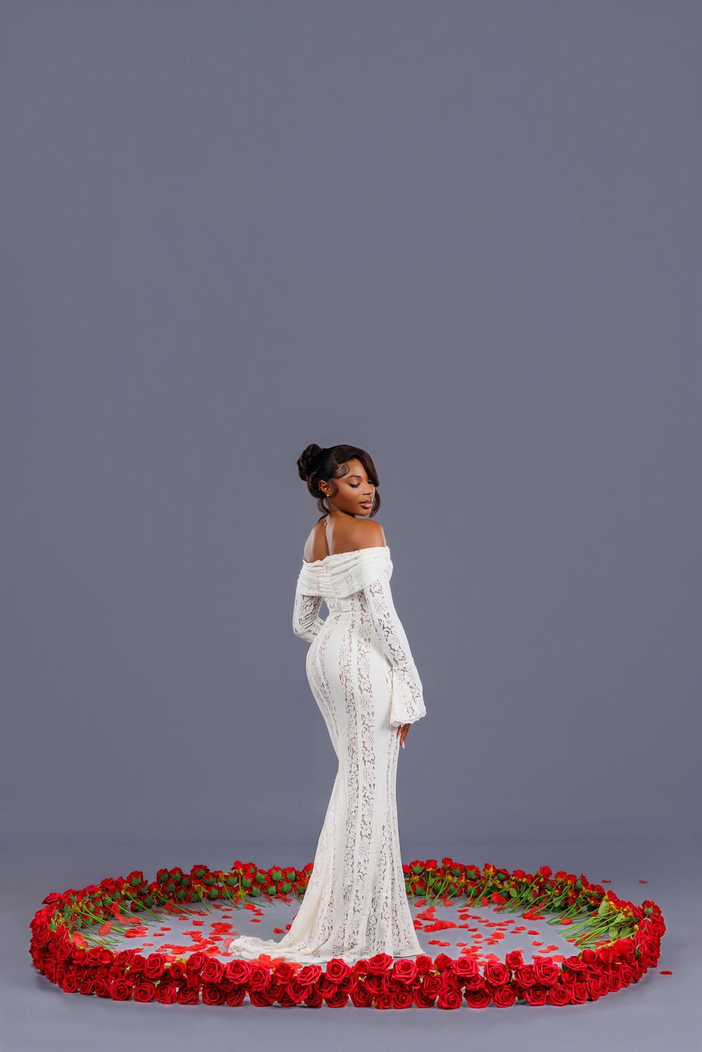 A bridal portrait session with a white lace gown surrounded by scattered red rose petals against a grey backdrop.