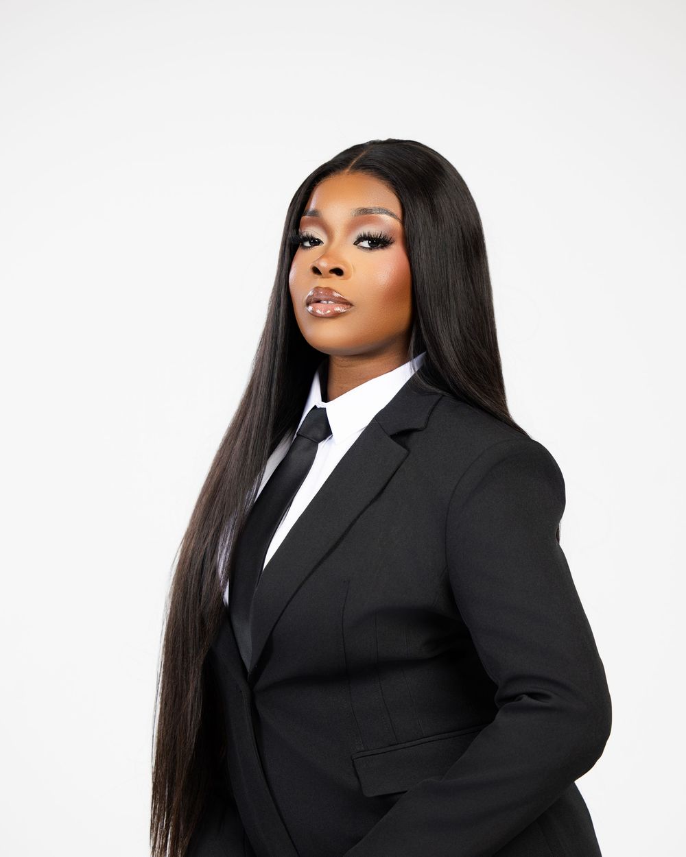 Professional portrait of someone in a black suit and tie with long straight dark hair against a white background.