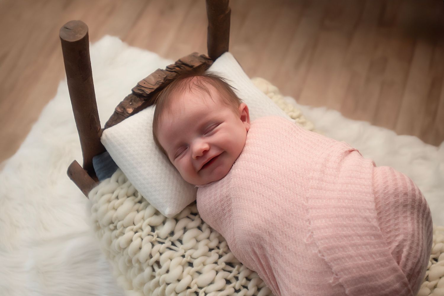 Baby wearing pink sweater lies on white textured carpet while reaching toward wooden furniture leg.