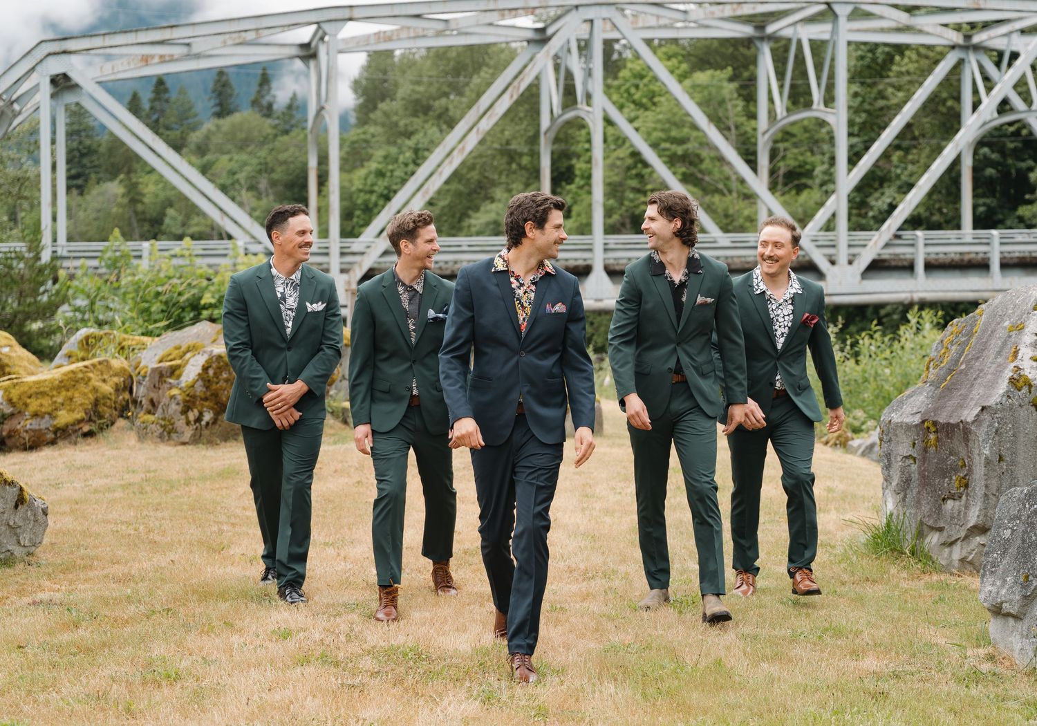 Wedding party in dark suits walks across a rustic bridge while laughing and enjoying a candid moment together.