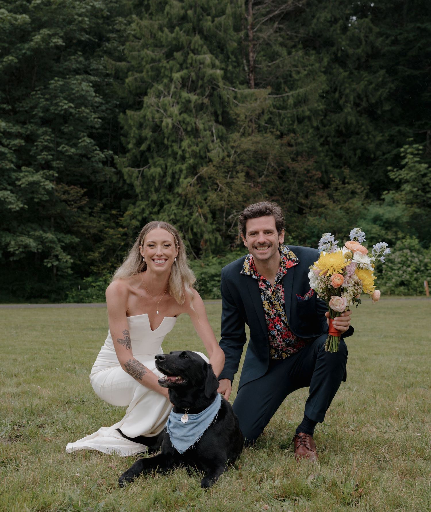 A playful wedding couple and their dog interact on a grassy lawn surrounded by trees.