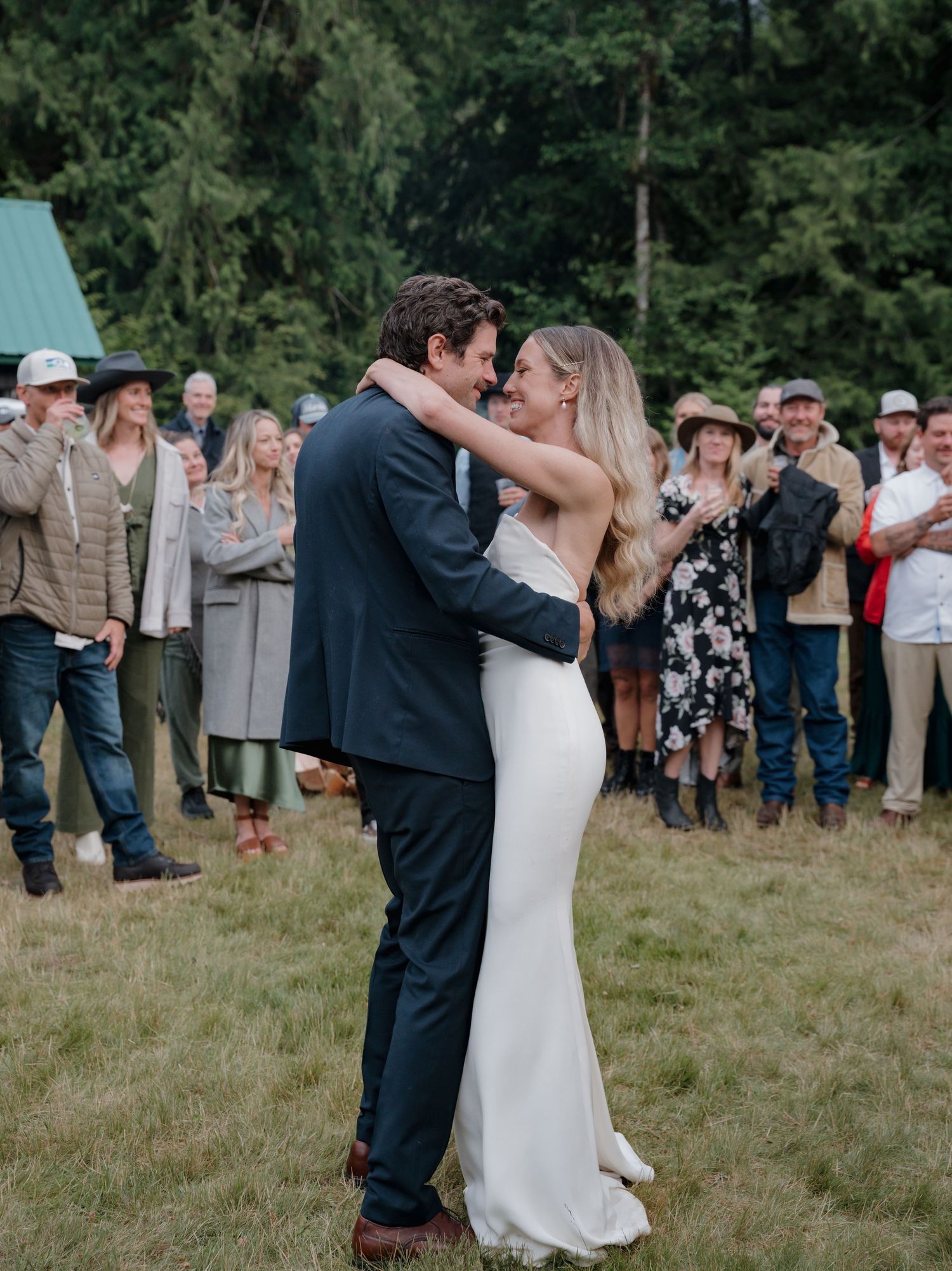 Couple shares their first dance as newlyweds while wedding guests watch from the background in an outdoor forest setting.
