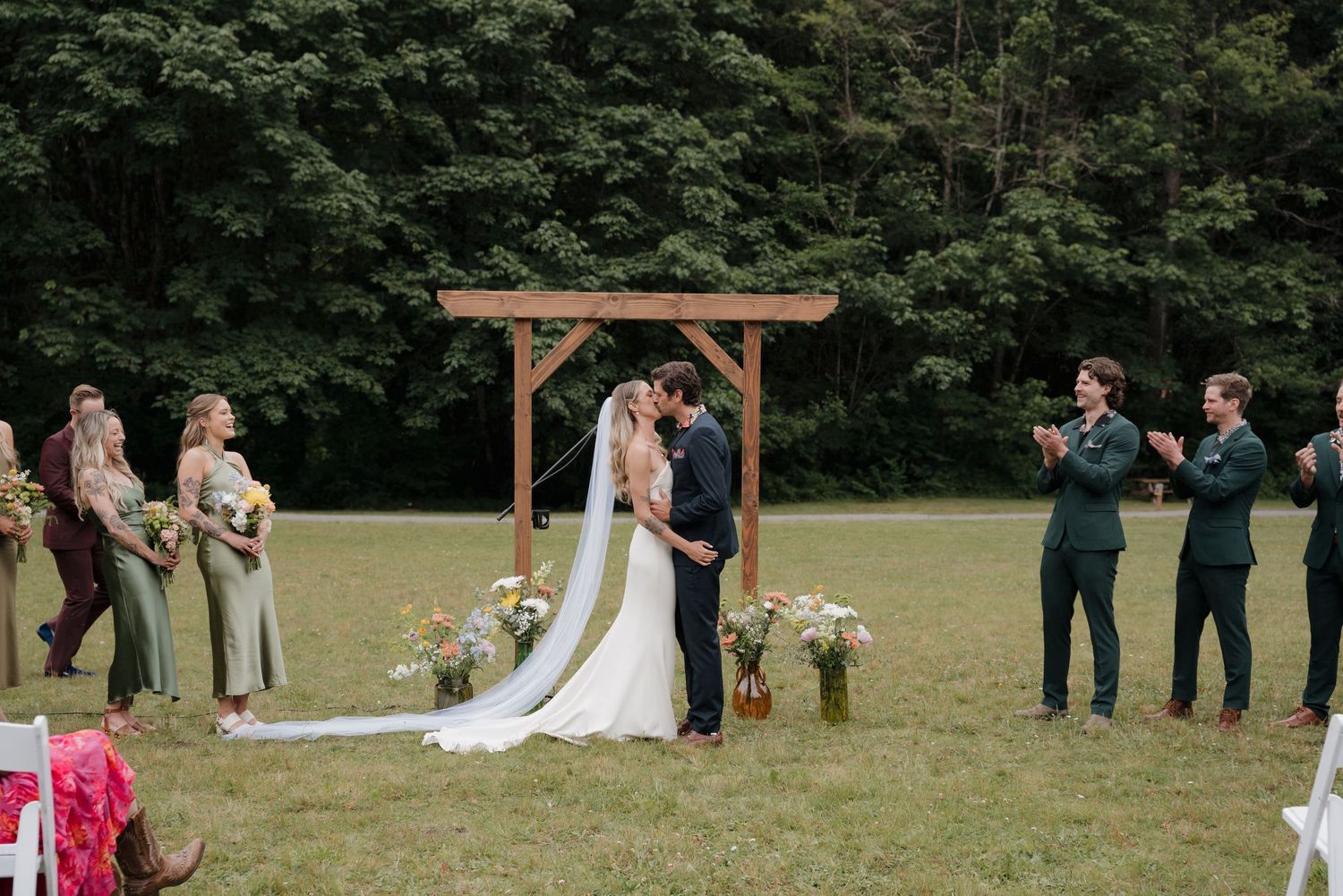 Wedding party stands in a line on a grassy field during an outdoor ceremony with trees in the background.