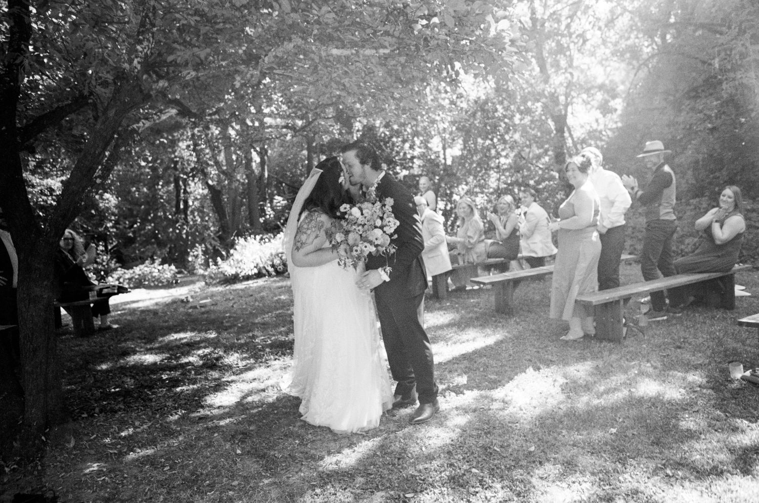 Black and white photo of a wedding celebration in a natural woodland setting with scattered sunlight.