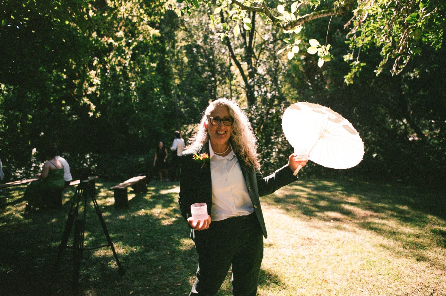 Wedding party walking through a sun-dappled garden path with parasols and flowing dresses.