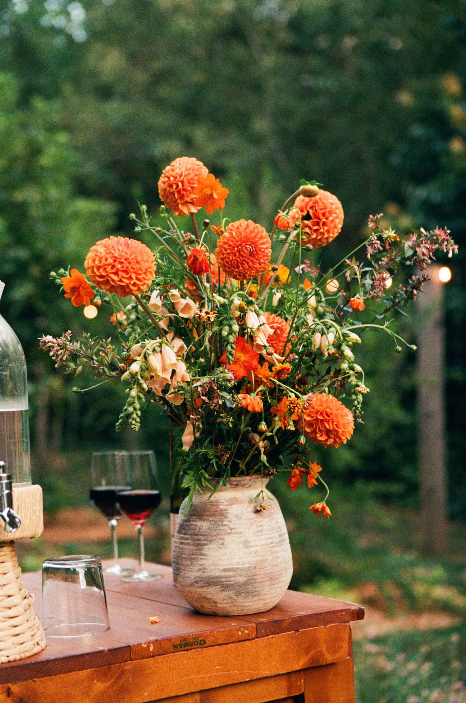 Orange dahlias in rustic vases displayed on wooden tables with wine glasses.