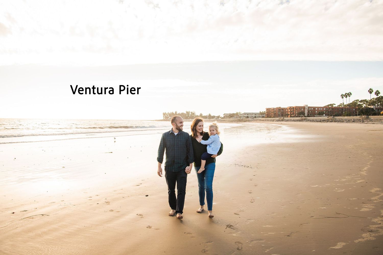 A couple walks along the sandy beach near Ventura Pier at sunset with palm trees in the background.