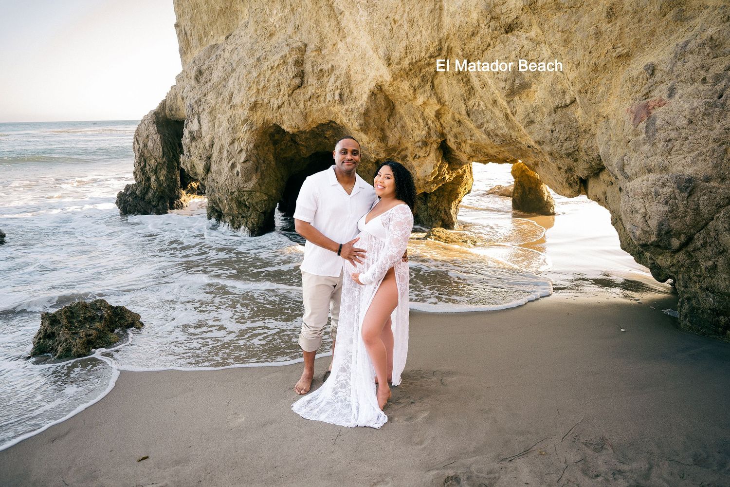 A romantic couple embraces on a scenic beach with rocky cliffs and crashing waves during golden hour.
