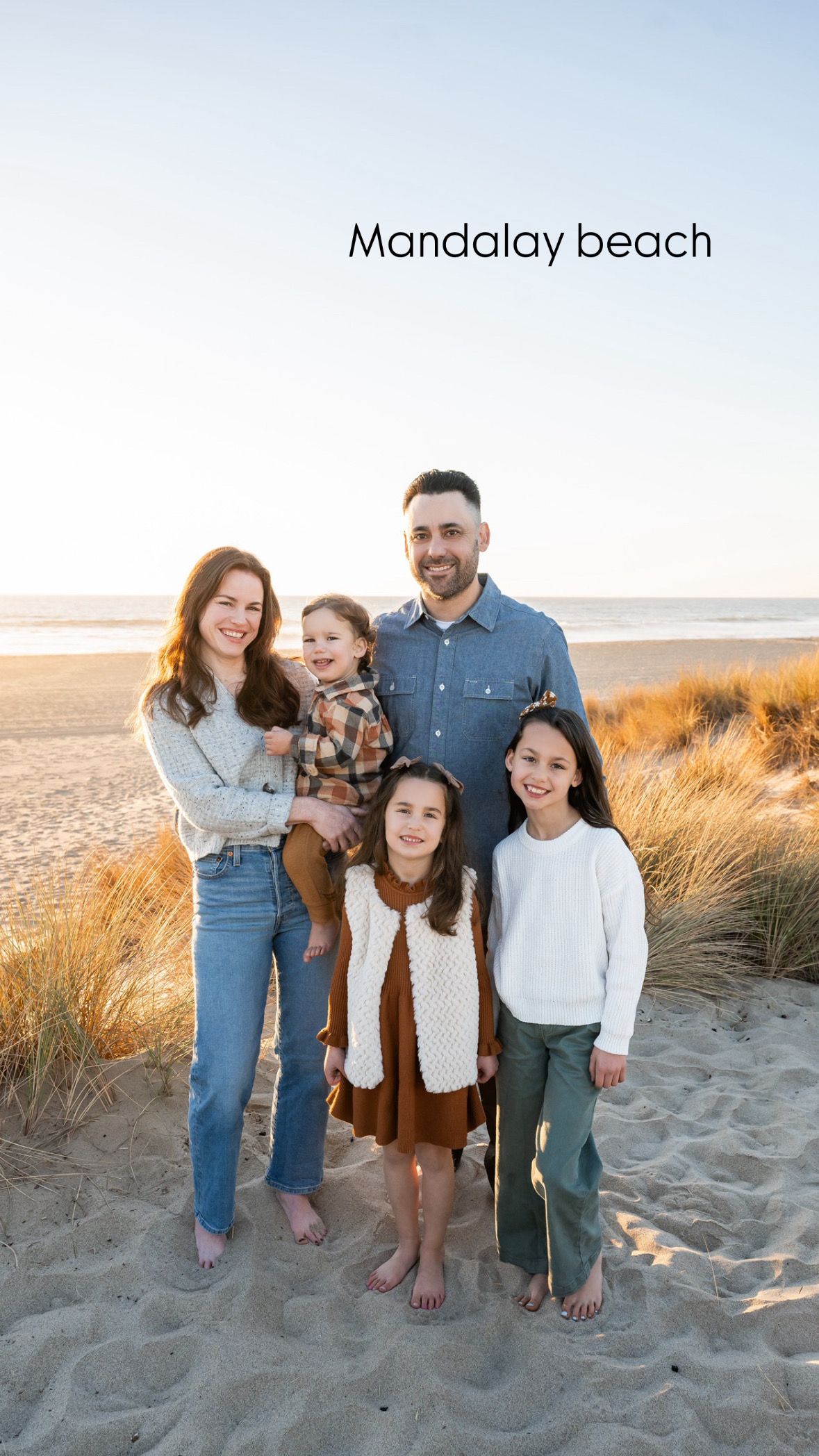 A family stands together on Mandalay Beach at sunset with coastal grass and sand dunes in the background.