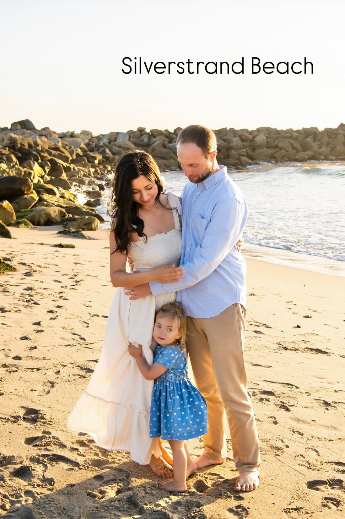 A family shares a tender moment together on Silverstrand Beach at sunset with rocky coastline in the background.