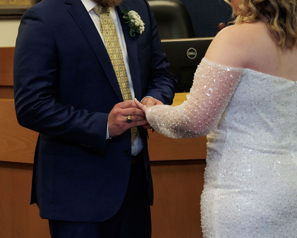 Close-up of hands exchanging wedding rings during ceremony with bride in sparkly white dress and groom in navy suit.