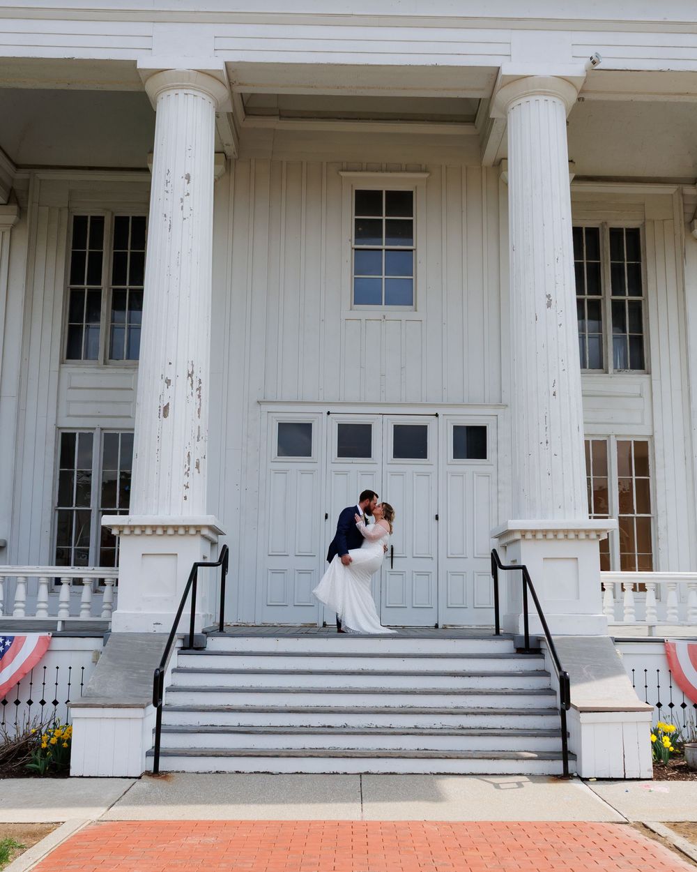 A wedding couple stands on the grand white steps of a neoclassical building with tall columns and ornate doorway.