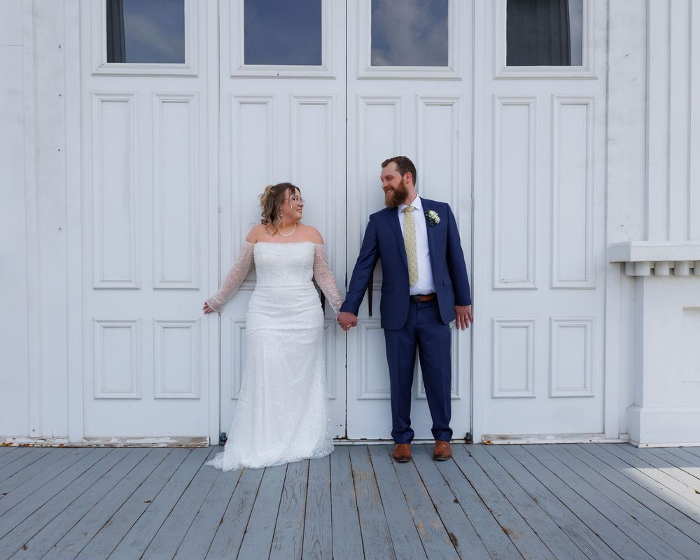 A bride and groom hold hands while standing on a white wood porch in front of tall white doors.