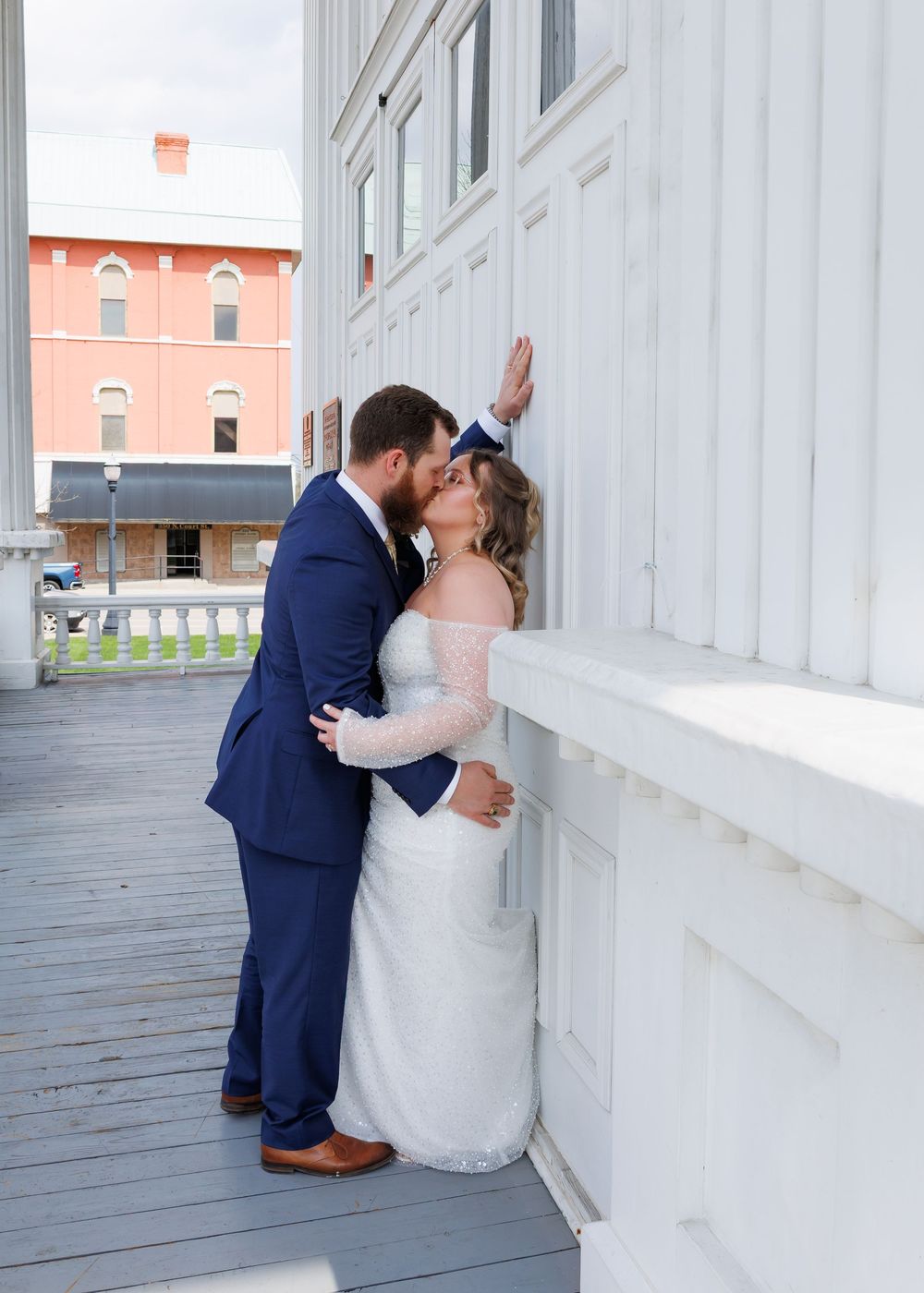 A romantic wedding couple shares an intimate moment on a white porch with a coral colored building in the background.