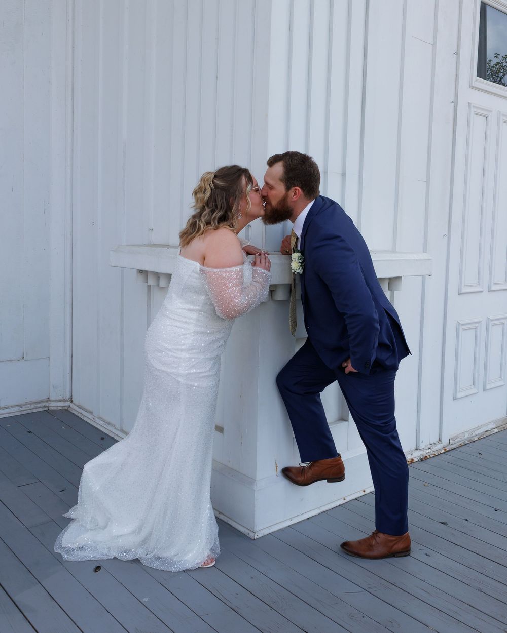 A couple shares a kiss on a white wooden porch, with the bride in a lace gown and groom in a navy suit.