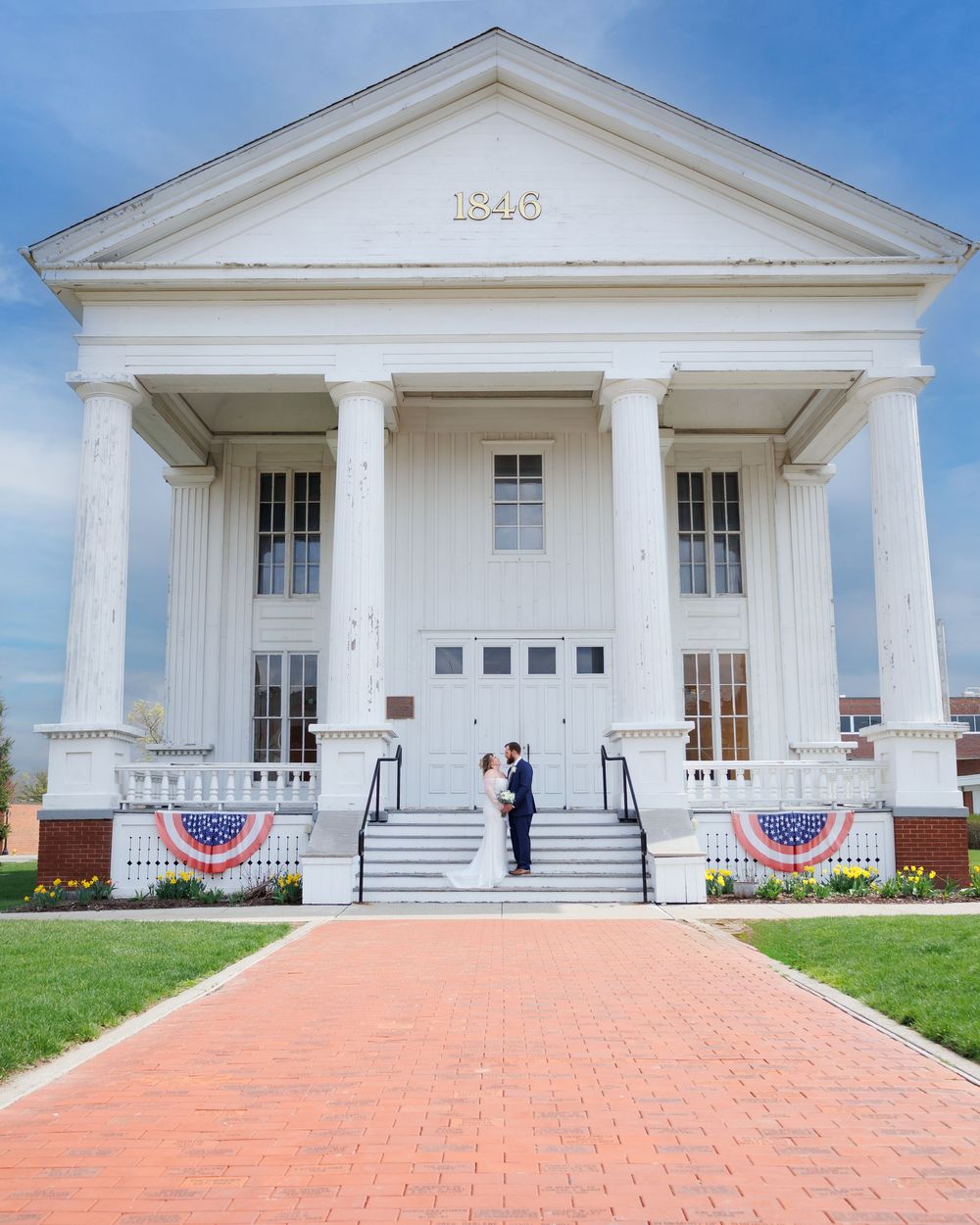 Historic white colonial building with columns and American bunting decorations, featuring a brick walkway leading to grand steps.
