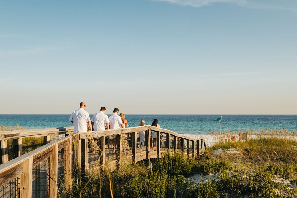 Panoramic view of people walking along a wooden boardwalk leading to a white sandy beach at sunset.