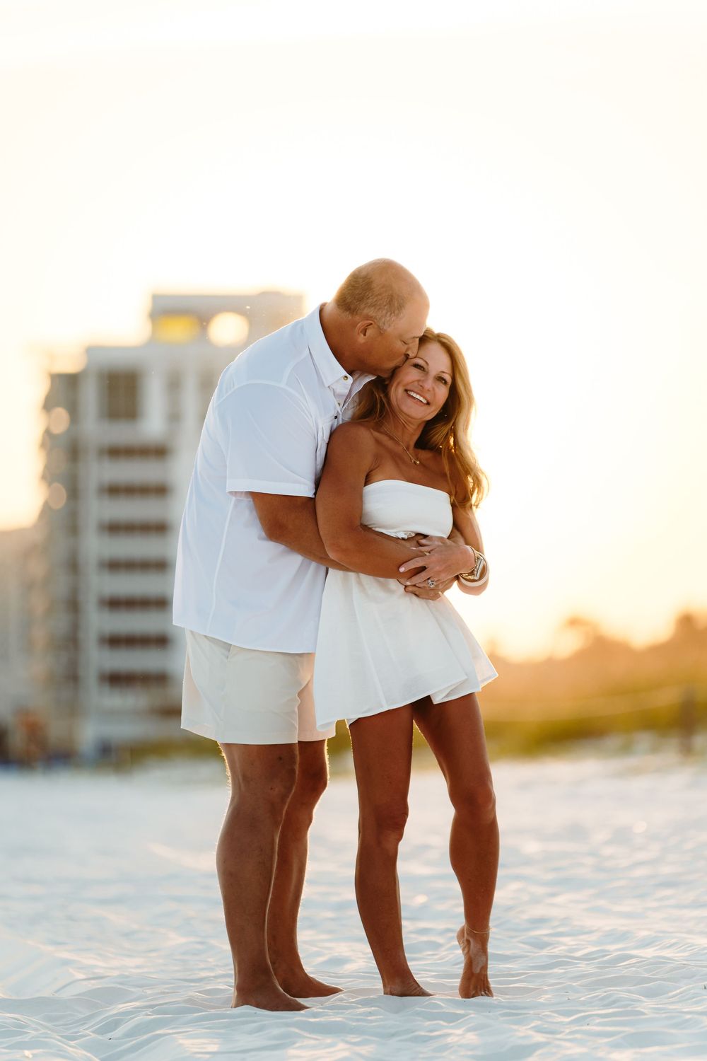 A romantic couple embraces on a white sandy beach at sunset with a coastal building in the background.