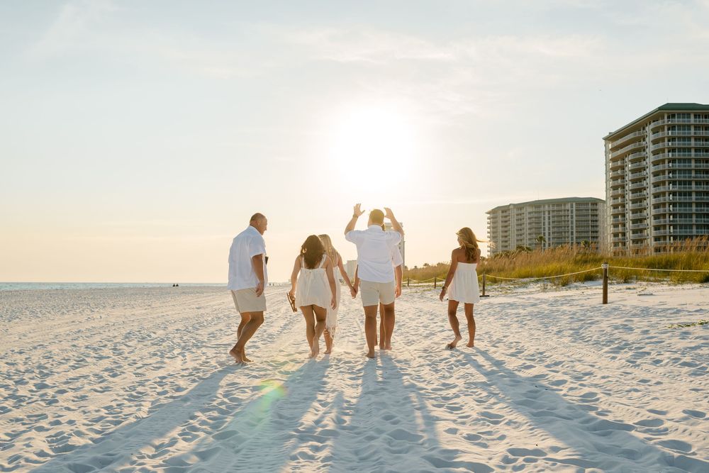 Family silhouettes walking together on pristine white sand beach with coastal buildings in background.