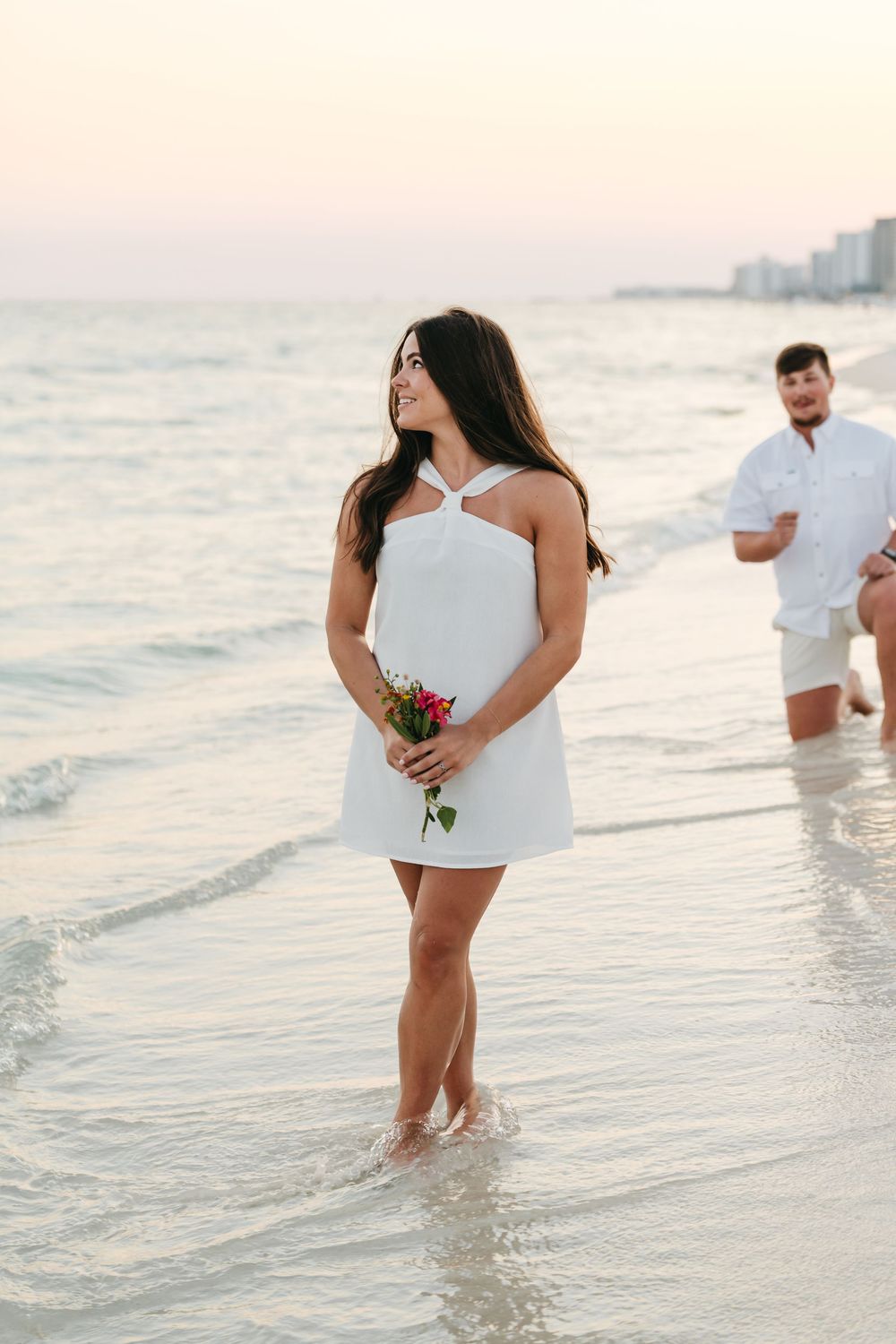 People in white clothing wade in shallow ocean water at sunset with beachfront buildings visible in the distance.